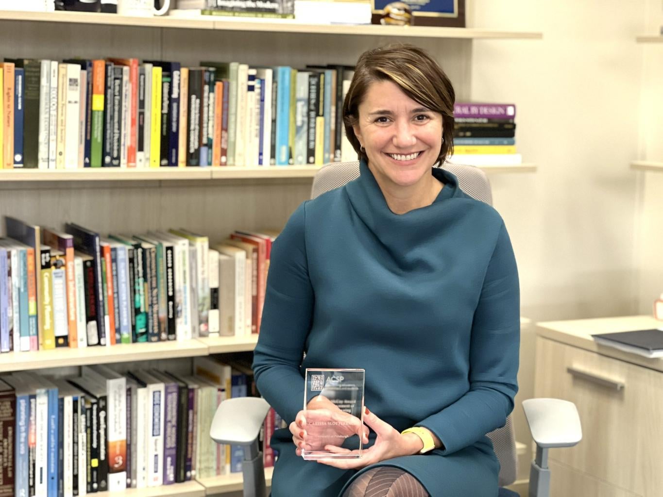 "Carissa Slotterback smiles, holding a glass award plaque. She sits in an office in front of a full bookshelf."