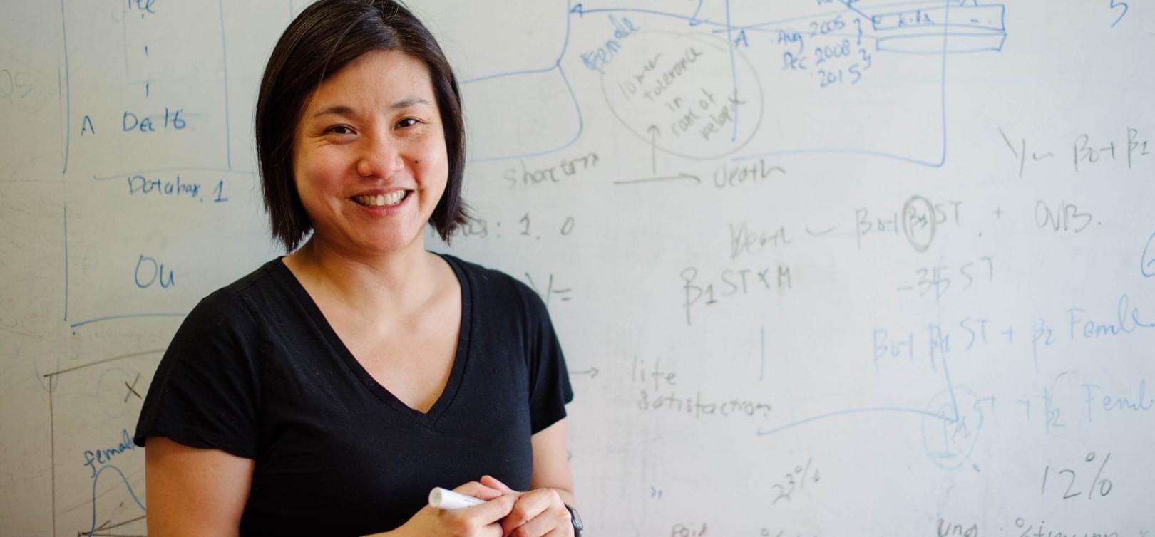 "Professor Sera Linardi holds a marker in front of a whiteboard full of graphs and equations"