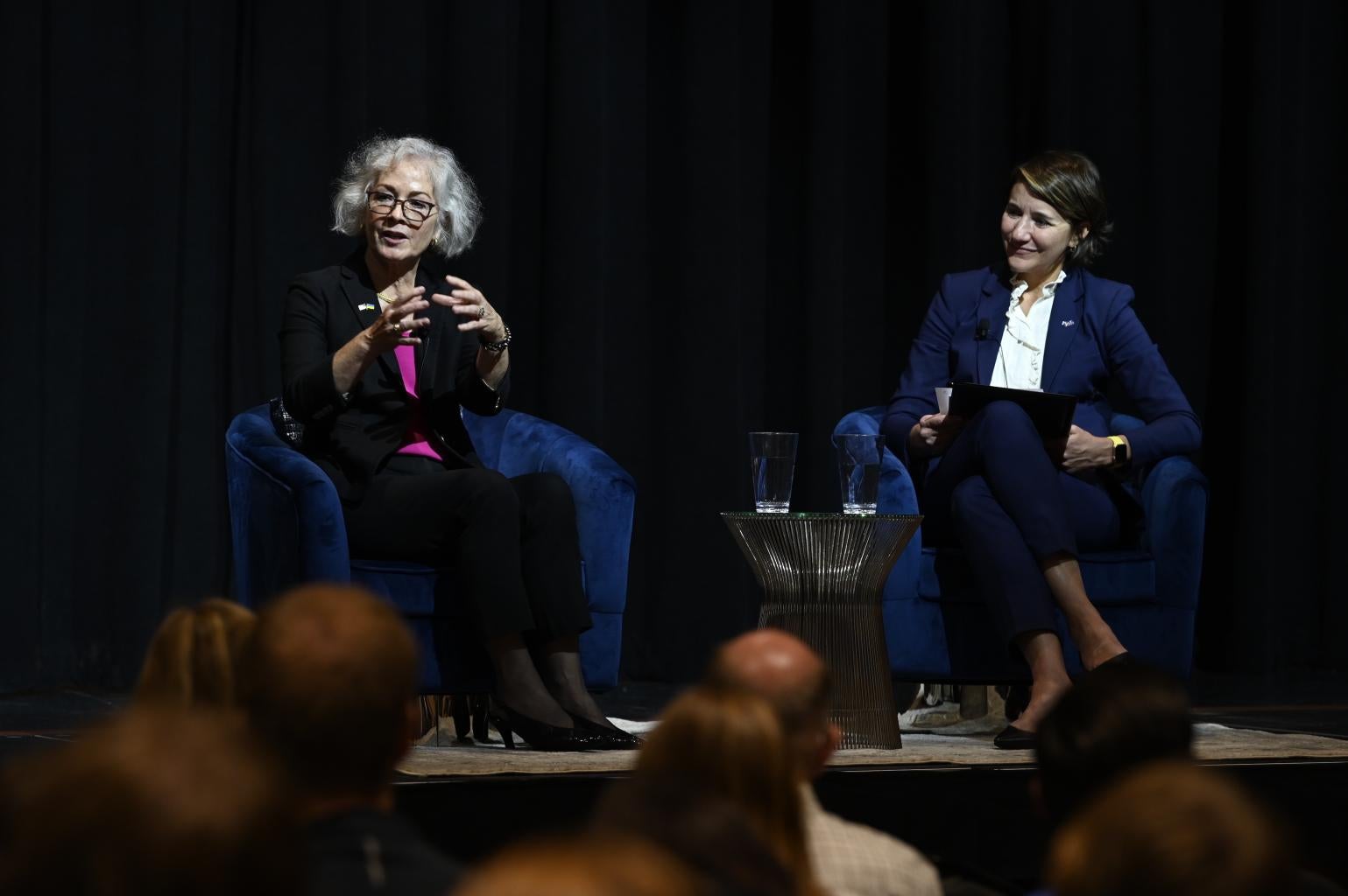 "Marie Yovanovitch and Carissa Slotterback sit opposite each other on a stage, talking. The tops of heads of people in the crowd are visible, facing the stage."