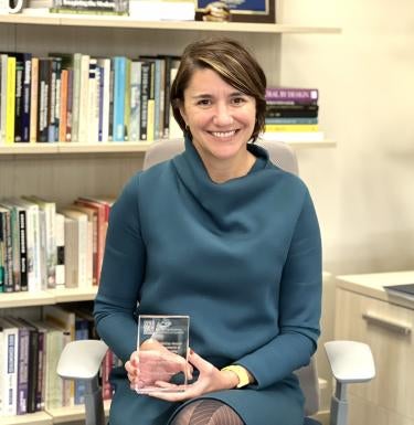 "Carissa Slotterback smiles, holding a glass award plaque. She sits in an office in front of a full bookshelf."