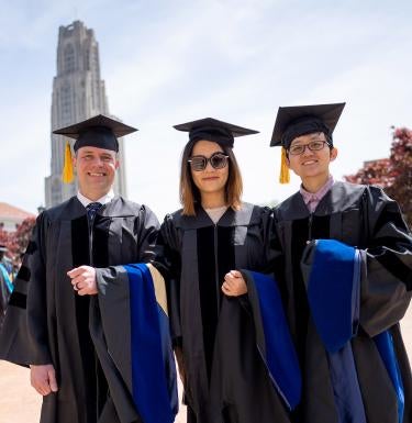 3 smiling people wearing graduation regalia stand together in front of the Cathedral of Learning.