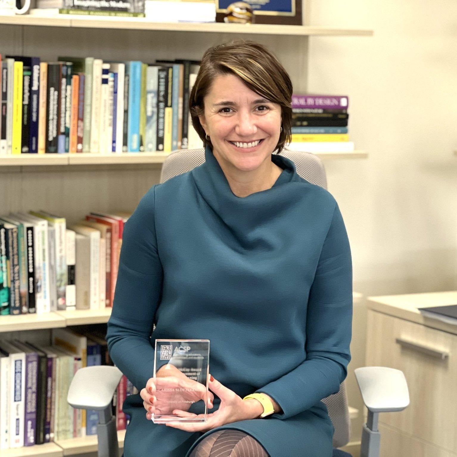 "Carissa Slotterback smiles, holding a glass award plaque. She sits in an office in front of a full bookshelf."